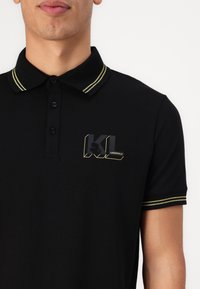 Čierne polo tričko s golierom so žltými pruhmi, štvoricou gombíkov a potlačeným logom na hrudi pre jedinečný dizajn.