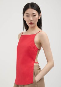 Top rosso senza maniche con spalline sottili, caratterizzato da un design asimmetrico e ritagli laterali. Realizzato in tessuto liscio con una leggera lucentezza.