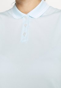 Ljusblå pikétröja med tre knappslå och ribbad krage som bärs av en person med synlig hals och haka.