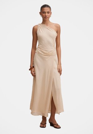 Vrouw die een beige maxi-jurk met één schouder draagt met dunne diagonale strepen, een zijsplit, sandalen en een bruine armband om haar pols.