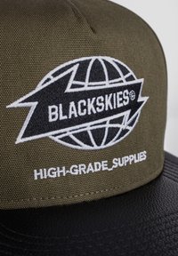 Gorra verde oliva hecha de tela texturizada con un visera de cuero negro. Presenta un logotipo bordado en blanco que dice "BLACKSKIES" y "HIGH-GRADE_SUPPLIES."
