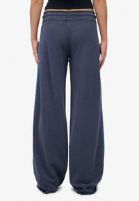 Persoon in losse donkerblauwe sweatpants met lichtblauwe strepen aan de zijkanten, staand tegen een effen witte achtergrond.