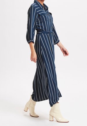 Námořnicky modrý pruhovaný overal s dlouhými rukávy, límečkem a páskem v pase. Nošený s krémovými lesklými kotníkovými botami.