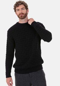 Schwarzer Strickpullover mit strukturiertem Webmuster, rundem Ausschnitt sowie gerippten Bündchen und Saum. Das Modell trägt graue Hosen.