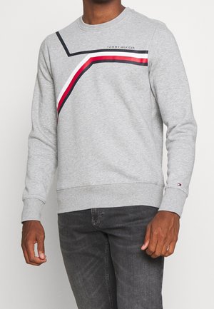 Mann trägt graues Tommy Hilfiger Sweatshirt mit roten, weißen und schwarzen schrägen Streifen über der Brust, kombiniert mit dunkelgrauen Jeans.