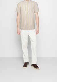 Chemise à manches courtes à boutons, beige avec des rayures verticales bleues et blanches. Associée à un pantalon blanc et des baskets marron foncé.