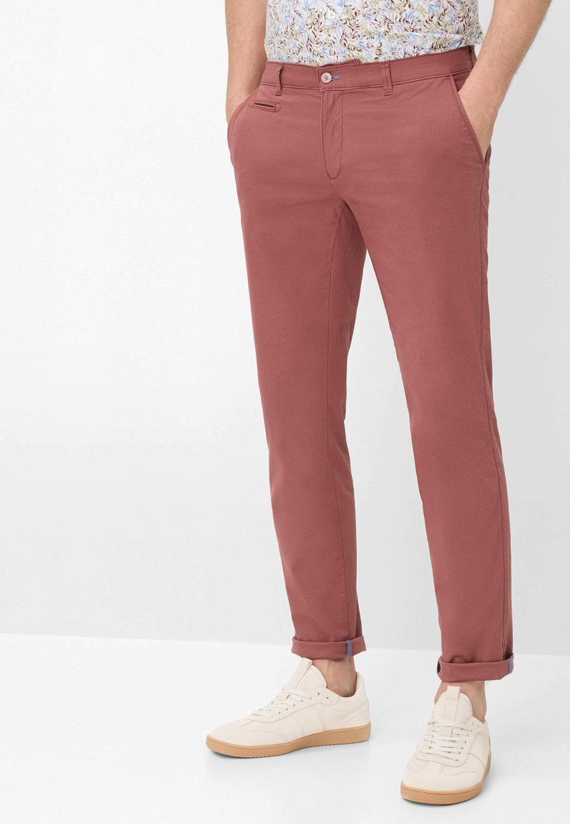 Homme portant un chino rose poudré retroussé, une chemise florale et des baskets écrues, debout les mains dans les poches sur un fond blanc.