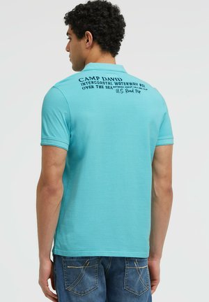 Mann trägt hellblaues Poloshirt mit schwarzem Text am oberen Rücken und blaue Jeans, steht rückwärts vor einfarbigem Hintergrund.