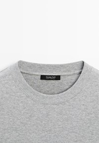 Gros plan sur le col d'un sweat-shirt gris à col rond avec une étiquette noire indiquant Massimo Dutti Made in Portugal.