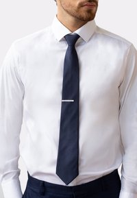 Cravate en soie bleue marine avec une texture lisse, ornée d'une fine barre d'accent argentée près de l'extrémité étroite, portée sur une chemise blanche.