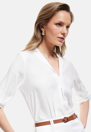 Blusa blanca con escote en V, tejido semitransparente y mangas enrolladas; combinada con un cinturón de cuero marrón con una hebilla redonda.