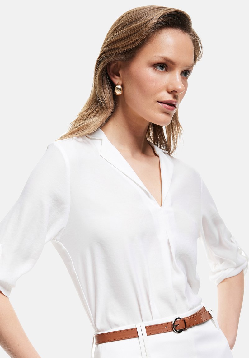 Blusa blanca con escote en V, tejido semitransparente y mangas enrolladas; combinada con un cinturón de cuero marrón con una hebilla redonda.