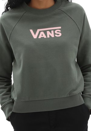 Personne portant un sweat-shirt Vans vert foncé avec un logo rose clair, mains dans les poches d’un pantalon noir, visible de la poitrine aux hanches.