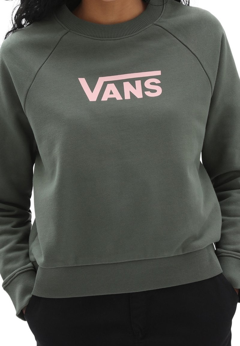 Personne portant un sweat-shirt Vans vert foncé avec un logo rose clair, mains dans les poches d’un pantalon noir, visible de la poitrine aux hanches.