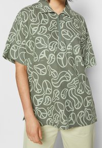 Camicia a maniche corte con colletto in verde oliva con motivo astratto di foglie verde chiaro. Presenta una zip frontale e una tasca applicata.