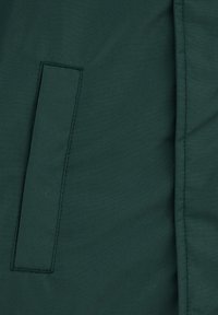 Tumši zaļš audums ar vertikālu taisnstūra šūtu kabatu un redzamu šuvi, kas iet pa labo pusi.