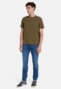 T-shirt verde oliva a maniche corte con colletto a girocollo, abbinata a jeans blu slim-fit e sneaker nere con strisce bianche.
