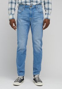 Jean bleu clair avec coupe droite, texture délavée, design à cinq poches et fermeture à bouton métallique, associés à des baskets montantes noires.