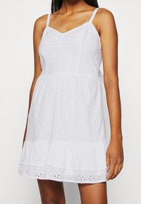 Vit ärmlös klänning tillverkad av bomull med ett ögla-mönster, volangkant och figurnära midja, med tunna axelband och V-ringning.