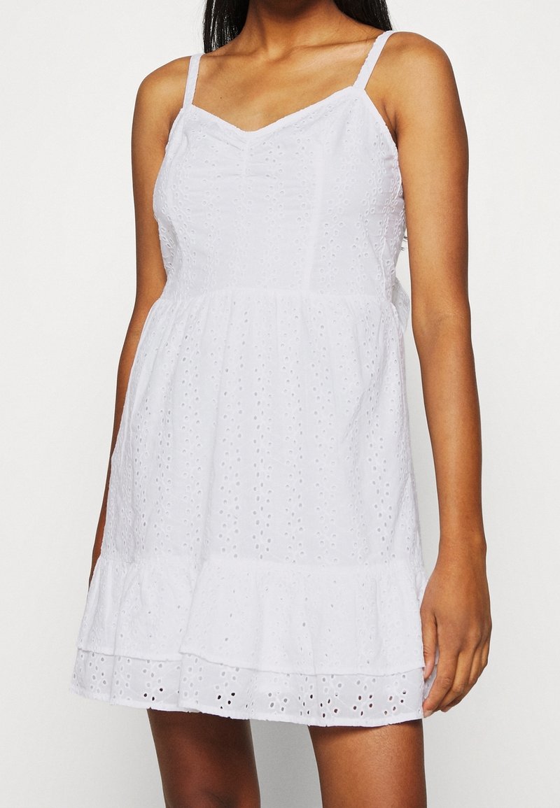 Vit ärmlös klänning tillverkad av bomull med ett ögla-mönster, volangkant och figurnära midja, med tunna axelband och V-ringning.