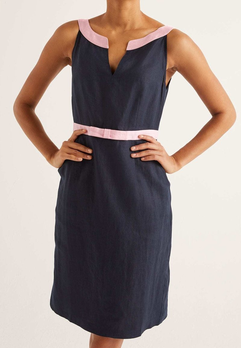 Femme portant une robe bleu marine sans manches avec une bordure rose pâle au niveau de l'encolure et de la taille, debout les mains sur les hanches.