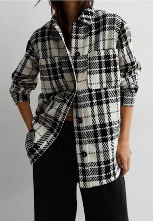 Femme portant une chemise à carreaux noire et blanche avec manches retroussées et pantalon noir, debout avec une main dans la poche.