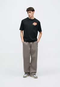 Fekete póló piros és sárga grafikai mintával, szürke melegítőnadrággal és szürke sportcipővel, amely fehér és fekete részleteket tartalmaz.