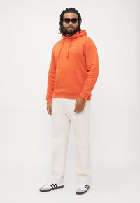 Uomo con dreadlocks che indossa occhiali da sole neri, felpa arancione, pantaloni bianchi e sneakers bianche con strisce nere, in piedi contro uno sfondo neutro.