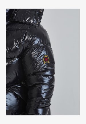 Blizgantis juodas pūkinis švarkas su gaubtu, su siuvinėtu raštu ir raudonu bei auksiniu logotipu kairiajame rankovėje.