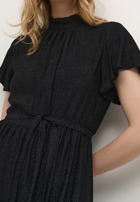 Černé šaty s krátkými volánkovými rukávy, vysokým výstřihem, staženým pasem a jemně třpytivou texturou. Rozšířená sukně s sklady.