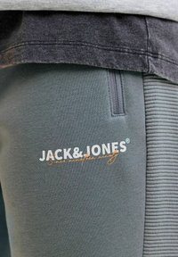 Grau-grüner Jogger aus strukturiertem Material, mit einer seitlichen Tasche und dem Logo "JACK&JONES" in Weiß und Orange. Weiches Material und gerippte Akzente.