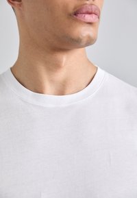 Tricou alb din bumbac, cu guler rotund, textură netedă și mâneci scurte, tăiat aproape pentru a evidenția detaliile gâtului și umărului.