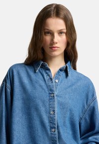 Camicia in denim di un azzurro chiaro con colletto, maniche lunghe e bottoni frontali. Presenta una vestibilità rilassata e oversize e una texture morbida.