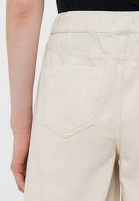 Lys beige bukser med elastisk talje, subtil tekstur, og en enkelt, diamantformet lomme på bagsiden.