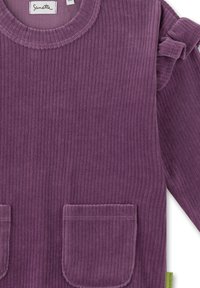 Lila gerippter Cordpullover mit zwei vorderen Taschen, rundem Ausschnitt und Rüschendetails an den Ärmeln. Ein Etikett an der Seite setzt einen kontrastierenden Akzent.