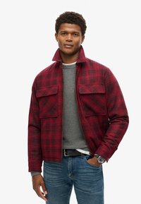 Chaqueta de cuadros roja y negra con un cuello alto, bolsillos en el pecho y cierre frontal de cremallera. Lleva puesta sobre un suéter gris texturizado y jeans azules.