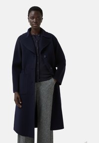 Navy wollen mixjas met grote kraag, knoopsluitingen en zijzakken, gecombineerd met een donkere trui en gestreepte broek.