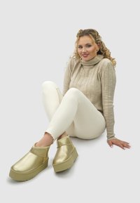 Gouden platformklompen gemaakt van glanzend materiaal met een hoge voorkant, ronde neus en gestructureerde rubberen zool. Gedragen met lichtgekleurde spijkerbroeken.