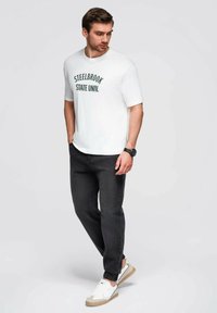 Camiseta blanca de manga corta con "STEELBROOK STATE UNIV." impreso en verde. Jeans oscuros y zapatillas blancas con acentos beige completan el look.