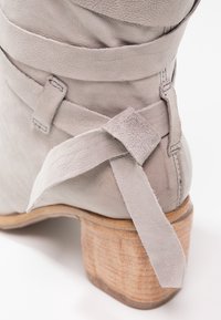 Bottine en cuir gris avec une finition texturée, détail de sangle enveloppante et un talon en bois empilé.