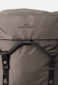 Sac à dos en nylon gris avec un rabat, accents noirs, fermeture à cordon de serrage et logo imprimé sur le devant. Détails en tissu texturé visibles.