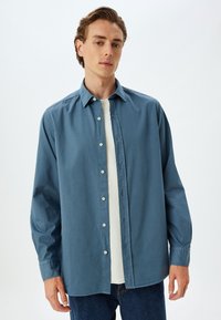 Camisa de botones azul claro, hecha de tela suave, con un cuello clásico, mangas largas y un corte relajado. Los botones son blancos.