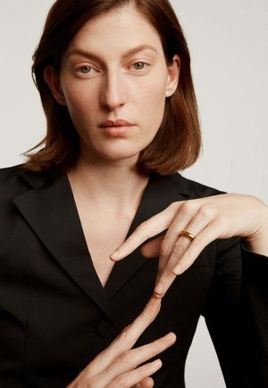 Anillo de oro en el dedo, modelo vistiendo una americana negra, manos desenfocadas, textura de piel suave, fondo claro.