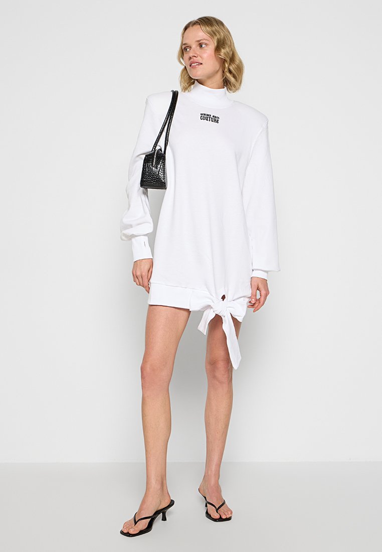 Blondynka ubrana w białą sukienkę mini z długim rękawem i węzłem z przodu, z czarną torebką na ramię i czarnymi sandałami na obcasie, stojąca przy białej ścianie.