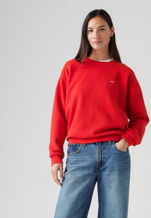 Roter Baumwollpullover mit rundem Halsausschnitt und gerippten Bündchen, kombiniert mit hellblauen Jeans. Verfügt über ein kleines Logo auf der Brust.