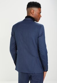 Mörkblå kostymjacka i texturerat tyg, slim fit, med en svart krage. Bakifrån syns strukturerade axlar och en split i fållen.