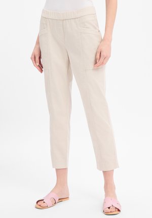 Beżowe krótkie spodnie z dużymi przednimi kieszeniami noszone z różowymi sandałami na białym tle.