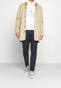 Cappotto lungo beige con bottoni, felpa bianca con logo, pantaloni a quadri blu navy e scarpe slip-on grigie. Design semplice con un aspetto casual.