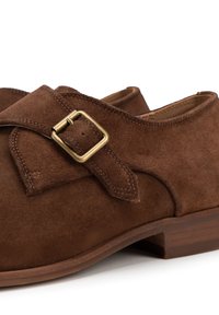 Zapatos de ante marrón con punta redondeada y una correa con una hebilla de latón. La suela es de madera con un tacón bajo.