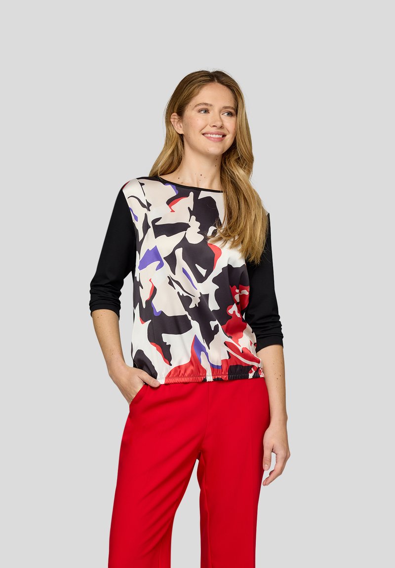 Langärmliges Oberteil mit einem schwarz-roten und weißen abstrakten Muster und einem elastischen Saum, kombiniert mit leuchtend roten Hosen, vor einem grauen Hintergrund.